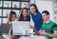 Vier junge, diverse Menschen sitzen lächelnd zusammen an einem Laptop im modernen Büro.