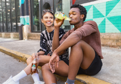 Ein junger Mann und eine junge Frau sitzen lachend auf einer Bordsteinkante und trinken Limonade.