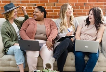 Vier gut gelaunte Frauen sitzen nebeneinander auf einem Sofa und arbeiten mit Laptops und Notizblöcken.