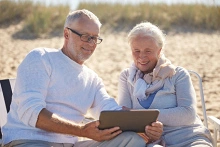 Ein älteres, lächelndes Ehepaar sitzt am Sandstrand und schaut gemeinsam auf ein Tablet.