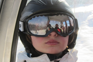 Nahaufnahme einer Influencer Person mit Skihelm, in deren Skibrille sich die Mitfahrer in einer Seilbahngondel spiegeln.